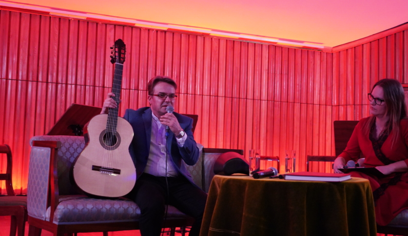 Mężczyzna siedząc przemawia do widowni, prawą dłonią przytrzymuje gitarę