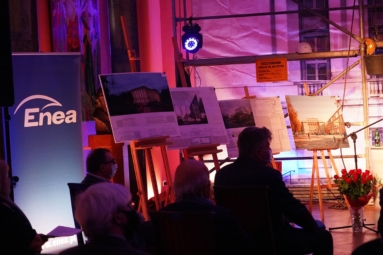 Grupa ludzi siedzi na krzesłach, w tle na sztalugach prace konkursowe