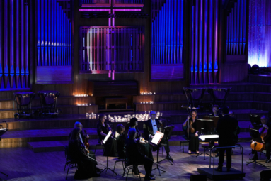 zdjęcie orkiestry kameralnej na przyciemnionej estradzie z wyświetlonym na prospekcie krzyżem