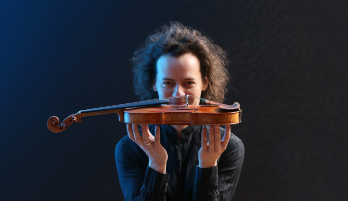 Mężczyzna z długimi włosami, trzyma przed sobą skrzypce