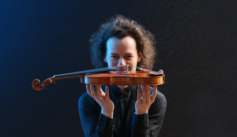 Mężczyzna z długimi włosami, trzyma przed sobą skrzypce