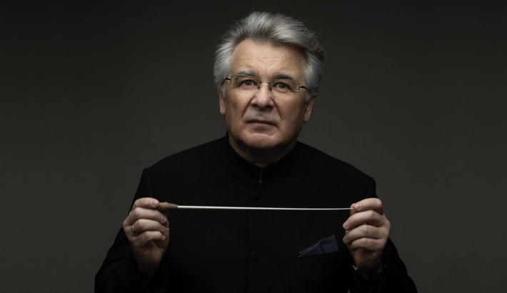 Mężczyzna z siwymi włosami, w okularach, trzyma przed sobą batutę