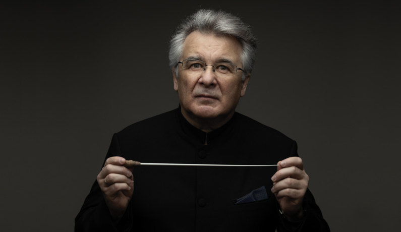 Mężczyzna z siwymi włosami, w okularach, trzyma przed sobą batutę