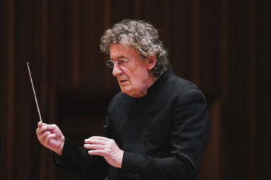 zdjęcie dyrygenta o siwych, kręconych włosach z okularami na nosie, w ciemnej marynarce