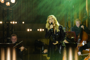 Kasia Cerekwicka ubrana w czarny, błyszczący kostium, śpiewa piosenkę trzymając w ręku mikrofon