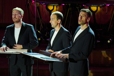 troje mężczyzn stoi przed pulpitami do nut i śpiewa utwór