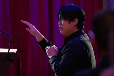 zdjęcie dyrygenta o azjatyckich rysach twarzy, z uniesioną prawą ręką