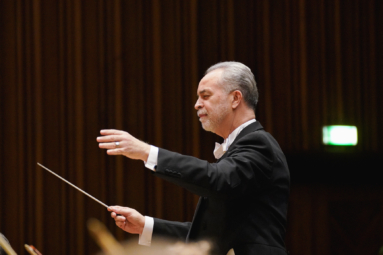 zdjęcie dyrygenta o siwych włosach i zaroście, z uniesionymi rękami i zamkniętymi oczami