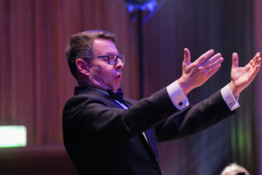 zdjęcie mężczyzny w garniturze i okularach z ciemnymi oprawkami, unoszącego ręcę w stronę chóru