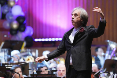 zdjęcie dyrygenta w siwych włosach z uniesioną ręką lewą, trzymającego batutę w ręce prawej