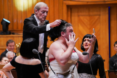 zdjęcie solisty mężczyzny przebranego w damski kostium oraz dyrygenta ściągającego soliście z głowy damską perukę