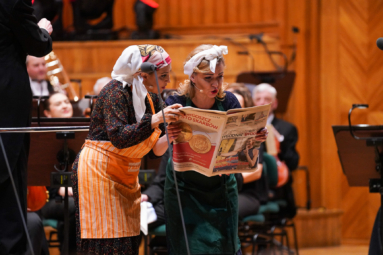 zdjęcie śpiewaczek ubranych w fartuchy kuchenne, z chustami na głowie, czytających gazetę