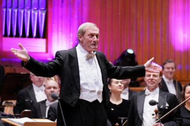 zdjęcie dyrygenta w starszym wieku, z siwymi włosami rozkładającego na boki ręce