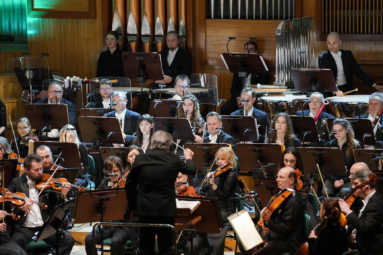 zdjęcie muzyków sekcji dętej orkiestry symfonicznej skupionych na utworze, czytających nuty leżące na puplitach przed nimi