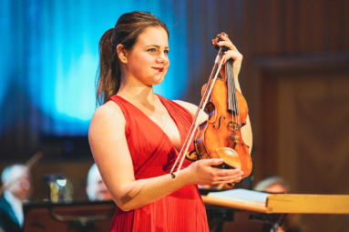 zdjęcie młodej dziewczyny w czerwonej sukience ze skrzypcami w rękach, uśmiechającej się w stronę publiczności