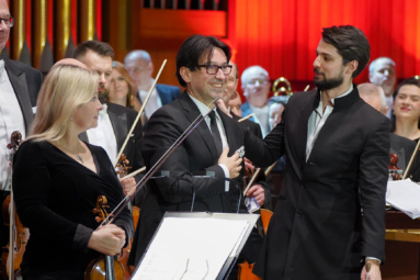 zdjecie mężczyzn w garniturach stojących na przodzie orkiestry. Jeden z nich spogląda na publiczność z ręką położoną na piersi, a drugi patrz na niego i wskazuje na niego ręką