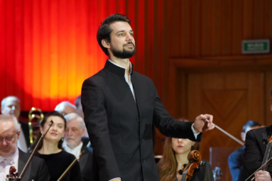 zdjęcie młodego mężczyzny w czarnych włosach stojącego na estradzie, trzymającego w ręku batutę, zwróconego w stronę publiczności