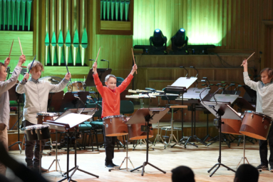 zdjęcie czwórki chłopczyków grających na instrumentach perkusyjnych, trzymających wysoko nad głową pałeczki perkusyjne