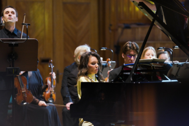 zdjęcie pianistki w żółtej sukience grającej na fortepianie, obok której stoi skrzypek z uniesioną ku górze głową