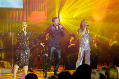 zdjęcie młodego mężczyzny i dwóch młodych kobiet w cekinowych strojach śpiewających do mikrofonów, w tle muzycy orkiestry kameralnej