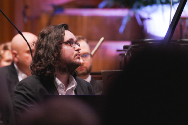 zdjęcie młodego mężczyzny w długich włosach i okularach grającego na fortepianie, wpatrującego się w dyrygenta