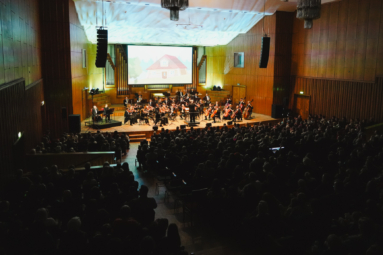 zdjęcie orkiestry symfonicznej na estradzie FP, nad którą wywieszony jest ekran multimedialny