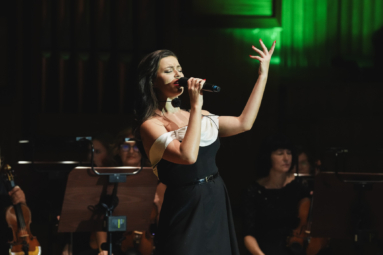 zdjęcie młodej kobiety śpiewającej do mikrofonu, unoszącej lewą rękę ku górze