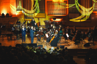 muzycy orkiestry kameralnej, zespołu i wokalistów na estradzie podświetlonej na kolorowo