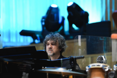 zdjęcie mężczyzny w w długich, lekko siwych włosach siedzącego za fortepianem