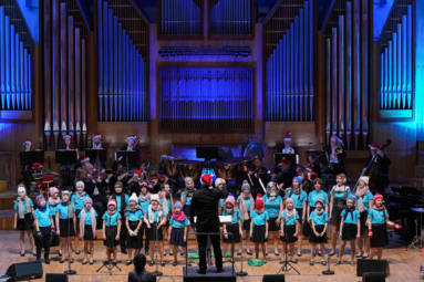 zdjęcie choru dziecięcego w niebieskich koszulkach wraz z orkiestrą kameralną na estradzie FP podczas koncertu