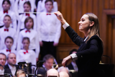 zdjęcie młodej dyrygentki w prostych ciemnych włosach uśmiechającej się w stronę orkiestry
