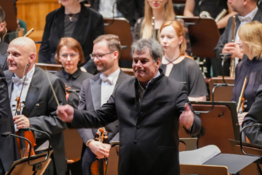 dyrygent z uniesionymi rękami, odwrócony i uśmiechający się do publiczności