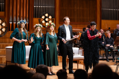 trzy kobiety w zielonych sukienkach, z wiankami na głowach oraz dwóch mężczyzn jeden we fraku, drugi w czerwono-czarnym przebraniu śpiewający w kwintecie