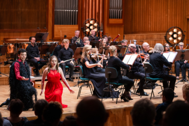 kobieta i mężczyzna śpiewający w duecie klęcząc na podłodze z towarzyszeniem orkiestry kameralnej