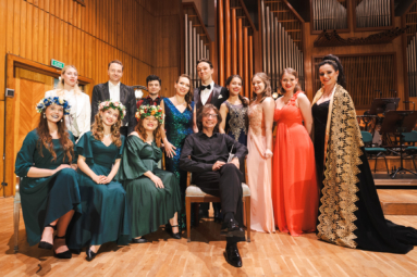 grupowe zdjęcie śpiewaków w estradowych strojach oraz dyrygenta siedzącego pośrodku nich na krześle