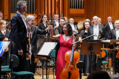 solistka podająca rękę dyrygentowi, a obok nich muzycy orkiestrowi z instrumentami w rękach