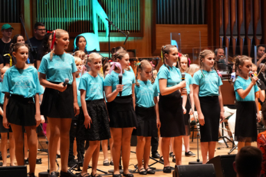 chór dziecięcy w niebieskich koszulkach i czarnych spódniczkach podczas kncertu na estradzie