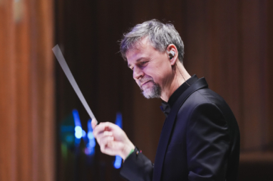 dyrygent z siwym zarostem w czarnej marynarce i słuchawce w uchu