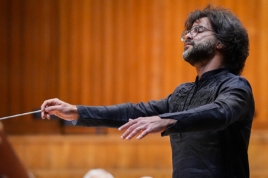 dyrygent w czarnych, bujnych włosach i czarnej koszuli, z rękami uniesionymi podczas dyrygowania