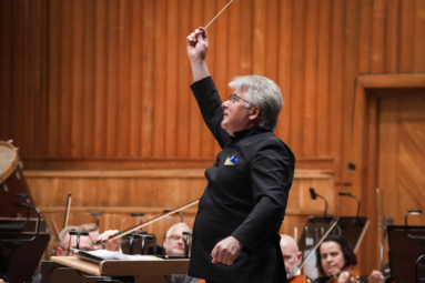 siwy dyrygent uśmiechający się z wysoko podniesioną batutą w prawej ręce