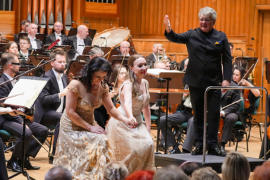 śpiewaczki kłaniające się do publiczności, a za nimi dyrygent wskazuje ręką w ich kierunku