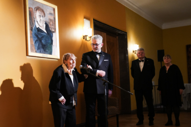 prof. Poniatowska przemawiająca przez mikrofon stojąc przed swoim portretem, podtrzymując za rękę dyrektora Puto