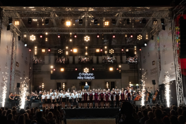 Na zdjęciu widoczna jest scena z chórem i orkiestrą podczas wieczornego koncertu, oświetlona licznymi reflektorami i efektami świetlnymi, a po bokach widać wybuchy sztucznych ogni,