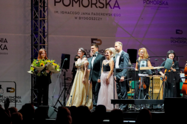 artyści na estradzie spoglądający w stronę publiczności, obok których stoi młoda dziewczyna z kwiatami