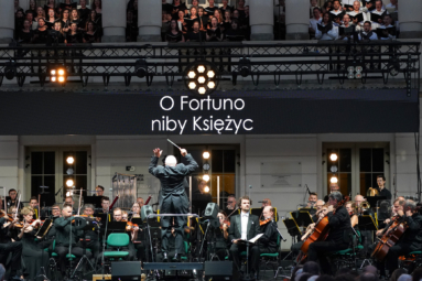 Zdjęcie przedstawia dyrygenta prowadzącego orkiestrę podczas koncertu plenerowego, nad którą wisi ekran z wyświetlonym napisem „O Fortuno niby Księżyc”, a w tle widoczny jest chór śpiewający z balkonu.