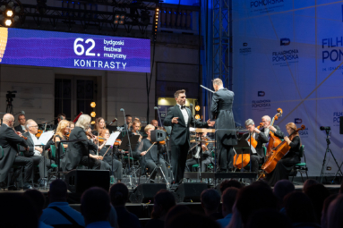 śpiewak w eleganckim garniturze i orkiestra kameralna na plenerowej estadzie, w tle napis 