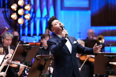 młody mężczyzna w granatowym garniturze śpiewający do mikrofonu z zamkniętymi oczami