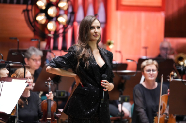 młoda kobieta w długich, prostych włosach, w jednej ręce trzyma mikrofon, a drugą podpiera na biodrze