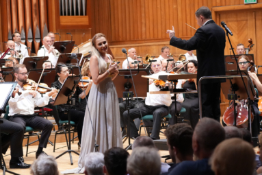 zdjęcie solistki w jasnej, długiej sukience uśmiechającej się do publiczności, przytulającej do siebie książkę, za solistką widoczni muzycy orkiestry