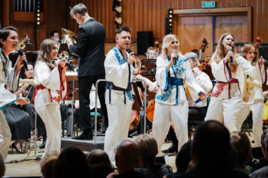 wokaliści w kolorowcyh strojach ABBY śpiewający do mikrofonów z towarzyszeniem orkiestry siedzącej za nimi